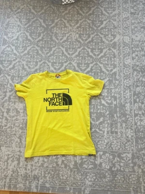 Gul The North Face t-shirt - Snygg gul t-shirt från The North Face med svart logga och text på bröstet. Klassisk rund halsringning och korta ärmar. Tillverkad i mjuk bomull som känns skön mot huden. Perfekt för dig som gillar streetwear och vill sticka ut med färg.