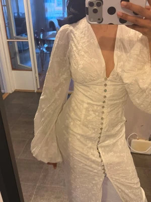 Adoore positano klänning <3 - Elegant vit långklänning med djup v-ringning, puffiga långärmar och dekorativa knappar framtill. Klänningen har broderade blommönster och en slits framtill för extra stil. Perfekt för dig som vill sticka ut med en romantisk look.