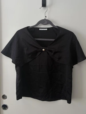 Svart satinblus med rosett - Vacker och elegant svart blus i satin/silkeslook  Fin detalj med stor rosett och pärlknapp framtill. Kort ärm och lätt, glansigt tyg.  Perfekt till både fest, middagar eller mer uppklädda tillfällen.  Super fin skick.  Storlek: Står ingen storlek, men skulle uppskatta en M, liten L     rosettblus bow blouse satin blouse festblus elegant top vintage style