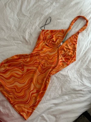 Orange mönstrad halterneck klänning - Säljer en kort halterneck klänning i orange och gula toner med ett vågigt, retroinspirerat mönster. Klänningen har smal axelrem och ärmlös design, perfekt för sommarens alla tillfällen. Materialet känns lätt och stretchigt.