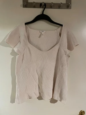 Ljusbeige ribbad topp från Object - Säljer en ljusbeige ribbad topp från Object med korta, fladdriga ärmar och djup rundad ringning. Toppen har en avslappnad passform och är tillverkad i ett mjukt, stickat material som ger en skön känsla. Perfekt att styla med jeans eller kjol.