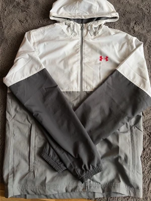 Gråvit vindjacka från Under Armour - Snygg vindjacka från Under Armour i grått och vitt med dragkedja framtill och huva. Jackan har elastiska muddar vid ärmsluten och två sidofickor. Under Armour-logga i rött på bröstet ger en sportig touch. Perfekt för blåsiga dagar.