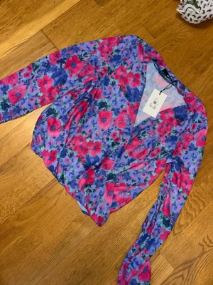 Blommig omlottblus från Bik bok  - Färgstark omlottblus från Bik bok med rosa och lila blommönster. Blusen har V-ringning och långa ärmar med lätt puff. Tillverkad i ett tunt, mjukt material som känns skönt mot huden. Perfekt för dig som vill sticka ut med en trendig look.