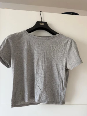 Grå ribbad croppad topp - En enkel och stilren grå croppad topp med ribbad struktur och korta ärmar. Perfekt till jeans eller kjol för en avslappnad look. Mjuk och skön mot huden.
