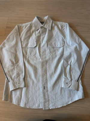 Vit skjorta med tryck på ryggen - Säljer en vit skjorta med coolt svart tryck på ryggen och kontrastsömmar längs ärmarna. Skjortan har två bröstfickor med knappar och klassisk krage. Perfekt för dig som vill sticka ut lite extra. Storleken är 146/152.