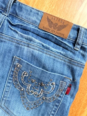 Bootcut jeans  med stenar från Botons - Snygga blå jeans från Botons med dekorativa nitar och broderi på bakfickan. Klassisk femficksmodell med normal midja och knappgylf. Jeansen har en cool tvättad look och detaljer i silverfärg. Midjemått tvärsöver: 37cm. Inre benlängd: 80cm. Bredd benöppning: 23cm 