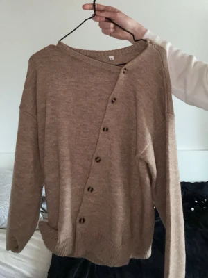 Beige kofta med sned knäppning - Säljer en beige stickad kofta med unik sned knäppning framtill och bruna knappar. Koftan har långa ärmar och ribbade muddar vid ärmslut och nederkant. Perfekt för lager på lager och enkel att styla till olika outfits.