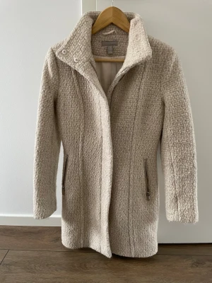 Beige kappa med hög krage - Säljer en beige kappa från H&M med två dragkedjefickor framtill. Jackan har en rak passform och stängs med dragkedja. 