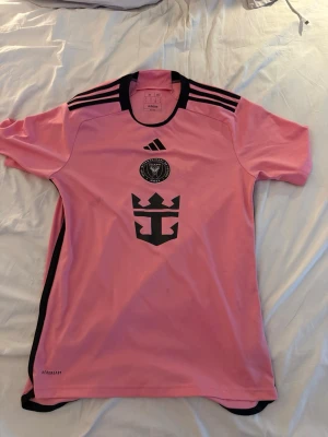 Rosa Inter Miami matchtröja Messi - Säljer en rosa Inter Miami matchtröja från Adidas med svarta detaljer och tryck. Tröjan har Messi och nummer 10 på ryggen, samt klubbmärke och sponsorlogga på framsidan. Tillverkad i lätt och ventilerande material med AEROREADY-teknologi.