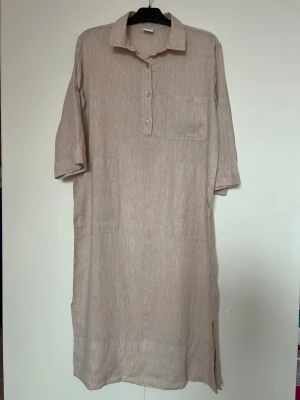 Ny, Beige linneskjortklänning med ficka i storlek S  - Stilren beige skjortklänning i linne med krage, knappar framtill och bröstficka. Halvlånga ärmar och slits i sidan ger en avslappnad look. Perfekt för varma dagar och enkel att styla med accessoarer.  Detaljer: 	•	Bröstficka 	•	Två fickor nedtill 	•	Ärmar som kan knäppas upp för kortare längd 	•	Slitsar i sidorna för ökad rörlighet  Storlek: S (36–38) Bredd:54cm Längd:117cm  Skick: Helt ny, aldrig använd Hem: Djur- och rökfritt hem