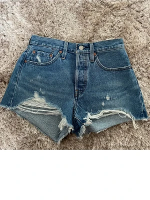 Levi's 501 blå denim shorts - Säljer ett par klassiska Levi's 501 shorts i blå denim med råa, fransiga kanter och slitningar framtill. Shorts med fem fickor, hög midja och knappar. Perfekt för dig som gillar en avslappnad och trendig look. Shortsen är oanvända med alla lappar kvar. Storleken är 26