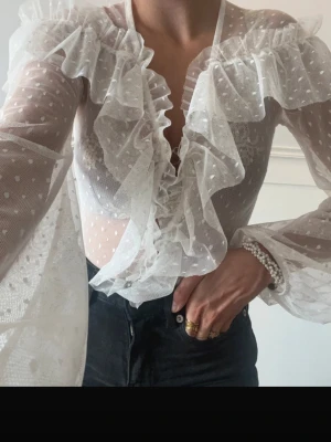 Vit transparent volangblus med prickar - Säljer en superfin vit blus i transparent mesh med små prickar och stora volanger längs urringningen. Blusen har långa ärmar och en romantisk, trendig look. Perfekt att styla med höga jeans eller kjol för en cool outfit.