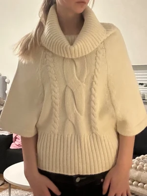 Vit stickad polotröja med flätmönster - Säljer en krämvit stickad tröja med stor polokrage och korta, vida ärmar. Tröjan har ett snyggt flätmönster framtill och bred ribbstickad kant nedtill. Perfekt för lager på lager och riktigt mysig till hösten och vintern. Storlek S