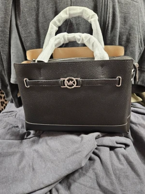 Svart handväska från Michael Kors - Snygg svart handväska från Michael Kors i strukturerat läder med silverfärgade detaljer och MK-logga framtill. Väskan har två handtag, dragkedja upptill och beige insida. Perfekt för dig som vill ha en stilren och trendig accessoar.