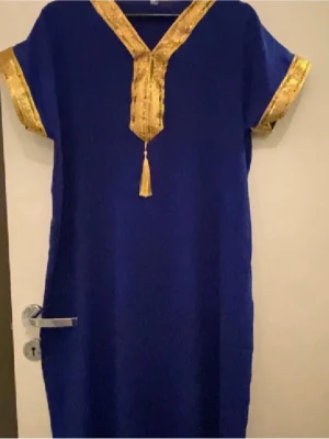 Blå klänning med guldiga detaljer - En blå klänning med raka linjer och korta ärmar. Klänningen har guldiga dekorband vid ärmslut och längs V-ringningen, samt en tofs framtill. Materialet ser ut att vara mjukt och lätt, perfekt för en unik och färgstark look.