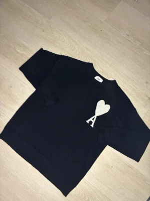 Svart stickad tröja från Ami Paris - Säljer en svart stickad tröja från Ami Paris med deras ikoniska vita hjärta och A på bröstet. Tröjan har rund halsringning, ribbade muddar och är gjord i mjukt material. Perfekt för dig som gillar stilrena och trendiga plagg.