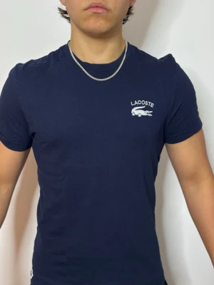 Mörkblå Lacoste t-shirt - Snygg mörkblå t-shirt från Lacoste med klassisk krokodillogo och text på bröstet. T-shirten har rund halsringning och korta ärmar. Tillverkad i mjuk bomull och har en slim passform som sitter tajt mot kroppen.