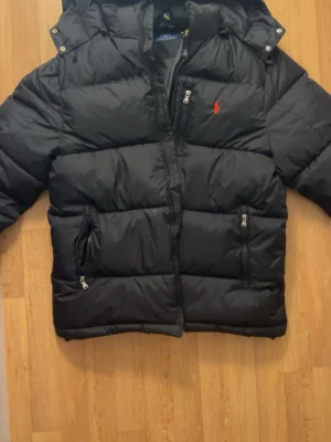 Svart dunjacka från Polo Ralph Lauren - Snygg svart dunjacka från Polo Ralph Lauren med huva och dragkedja framtill. Jackan har två framfickor med dragkedja och den klassiska röda loggan broderad på bröstet. Perfekt för kalla vinterdagar och riktigt stilren look.