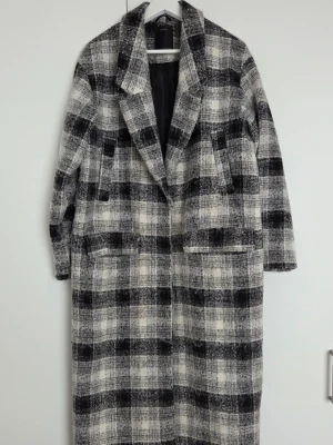 Lång rutig kappa - Lång kappa i svartvitt rutigt mönster. Kappan har breda slag, två stora fickor framtill och är fodrad. Använd men inga skador/fel. 
