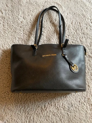 Svart handväska från Michael Kors - Stilren svart handväska från Michael Kors i strukturerat läder med dubbla axelband och guldfärgade detaljer. Väskan har ett dekorativt MK-hänge och logga i guld på framsidan. Perfekt för dig som gillar klassisk och tidlös design.