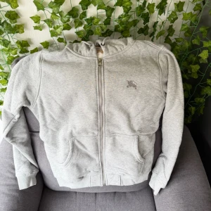 Grå zip hoodie från Burberry - Snygg grå hoodie med dragkedja från Burberry. Klassisk logga broderad på bröstet och ikoniskt rutigt Burberry-mönster på insidan av huvan. Två fickor fram och ribbade muddar. Perfekt för en chill och stilren look.