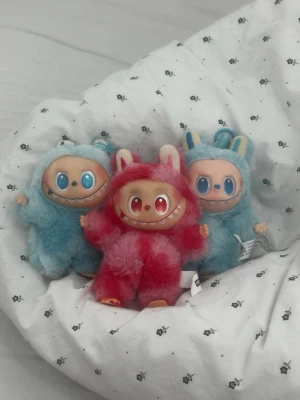 Tre fluffiga monster-gosedjur labubus - Säljer tre gulliga monster-gosedjur med stora leenden och färgglada pälsdräkter. Två är ljusblå med blå detaljer och kaninöron, en är röd/rosa med vita öron. Alla har stora glittriga ögon och mjuk päls. Perfekta som dekoration eller samlarobjekt.