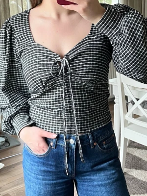 Svartvit topp med puffärm - Svartvit topp med rutigt mönster, puffiga ärmar och snygg knytning framtill du kan knyta så hårt/löst du vill. V-ringning och figurnära passform. Kan användas till vardag och fest. Passar M-XL skulle jag säga.