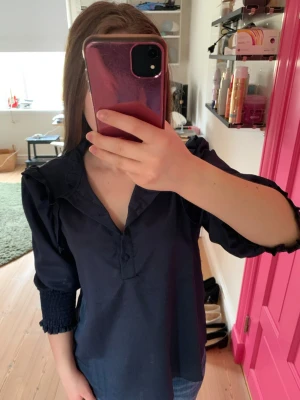 Mörkblå blus med volangdetaljer - Trendig mörkblå blus med volangdetaljer vid axlarna och knappar framtill. Blusen har trekvartsärm med smock vid ärmslut och en lätt upphöjd krage. Perfekt för dig som gillar en stilren men ändå lekfull look.