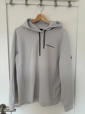 Ljusgrå hoodie från Peak Performance - Snygg ljusgrå hoodie från Peak Performance med svart dragsko i huvan och diskret logga på bröstet. Tillverkad i mjukt material och har en liten logga på ärmen. Perfekt för en avslappnad och stilren look.