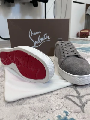 Gråa Christian Louboutin sneakers - Snygga gråa sneakers från Christian Louboutin i mocka med klassisk röd sula och vita kanter. Skorna har lågt skaft, matchande gråa skosnören och stilren design som passar till det mesta. Perfekta för dig som vill ha något exklusivt och trendigt.