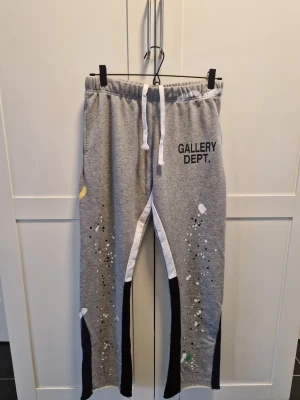 Grå mjukisbyxor från Gallery Dept. - Säljer ett par grå mjukisbyxor från Gallery Dept. med vita och svarta detaljer längs benen och färgstänk som ger en unik look. Byxorna har resår i midjan med vit dragsko och tryckt logga på vänster sida. Tillverkade i mjukt bomullsmaterial för maximal komfort.