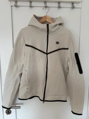 Beige Nike hoodie - Snygg beige hoodie från Nike med svart dragkedja och detaljer. Hoodien har huva, två sidofickor och en ficka med dragkedja på ärmen. Sportig design med svarta linjer över bröstet och Nike-logga på vänster sida. Perfekt för dig som gillar streetwear.