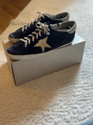 Mörkblå Golden Goose sneakers - Snygga mörkblå sneakers från Golden Goose med vit stjärndetalj på sidan och vita skosnören. Skorna har en låg profil och är tillverkade i mocka med vita detaljer på häl och sula. Perfekta för en trendig och avslappnad stil. Box ingår. Nypris: 5000 || Mitt pris: 1700