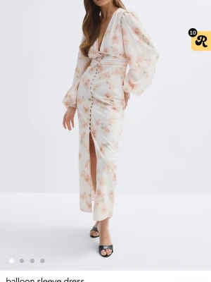 Lång blommig klänning med slits - Elegant långklänning i ljus bas med blommigt mönster i rosa toner. Klänningen har v-ringning, ballongärmar, knappar framtill och en slits. Tillverkad i ett lätt material som ger ett luftigt intryck. Perfekt för sommarens tillfällen.