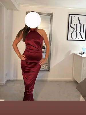 Vinröd långklänning i satin - Snygg vinröd långklänning i satin med halterneck och figurnära passform. Klänningen har en elegant glansig yta och är ärmlös, perfekt för speciella tillfällen. Stilren och modern design som framhäver siluetten.