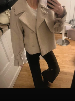 Beige ullrock med bred krage - Kort beige ullrock från H&M med bred krage och dubbelknäppning. Rocken har markerade axlar, vida ärmar med dekorativa spännen vid handlederna och en avslappnad passform. Perfekt för dig som vill ha en trendig och stilren look.