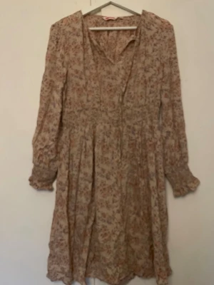 Blommig beige klänning med volang - Söt rosa/beige klänning med blommigt mönster och volangdetaljer. Klänningen har långärmade ärmar med resår vid ärmslut och en lätt v-ringad halsringning. Perfekt för vår och sommar, i ett luftigt material. Stl 42 men mer som en 38-40