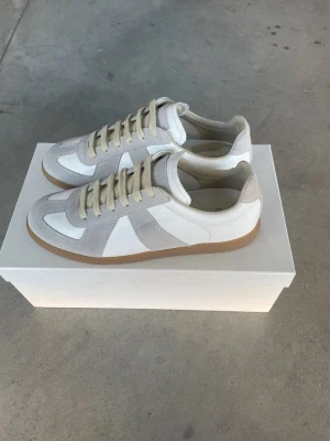 Vita Maison margiela gats - Snygga sneakers i vitt och ljusgrått med beige skosnören och klassisk gummisula. Ovandelen är i en mix av slätt läder och mocka för en stilren look. Perfekta för dig som gillar minimalistisk och clean stil.