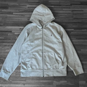 Grå hoodie från Polo Ralph Lauren - Snygg grå kofta från Polo Ralph Lauren med dragkedja framtill och två stora fickor. Klassisk broderad logga på bröstet. Tillverkad i mjukt bomullsmaterial och har en stor huva samt ribbade muddar vid ärmar och nederkant. (Saknar snören)