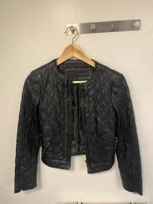 Svart quiltad skinnjacka Zara Basic - Snygg svart skinnjacka från Zara Basic med quiltat mönster och rundad krage. Jackan har dragkedjor vid ärmsluten och framtill, samt två fickor med dragkedja. Kortare modell som ger en trendig look.