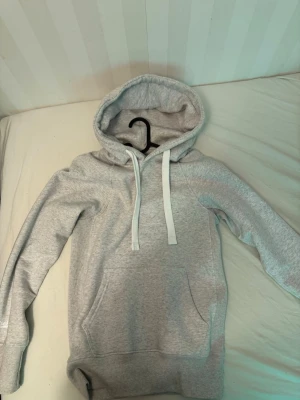 Ljusgrå hoodie från Russemerch - Ljusgrå hoodie från Russemerch med ficka framtill och vita dragsnören i huvan. Mjuk bomullsblandning och ribbade muddar vid ärmslut och nederkant. Diskret Russemerch-logga på ena ärmen. Perfekt för chill dagar.