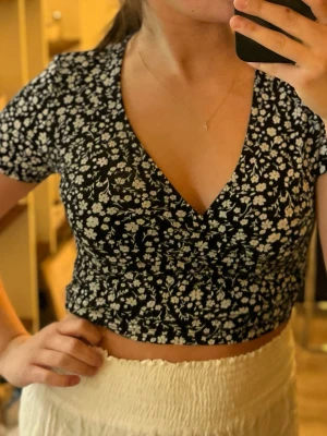Två omlottoppar med blommigt mönster - Säljer två omlottoppar i croppad modell, en svart med vita blommor och en vit med ljusblå blommor. Båda har korta ärmar, djup V-ringning och stretchigt material. En S & en M