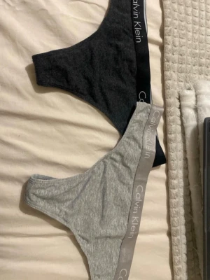 Calvin Klein stringtrosor i bomull - Två par stringtrosor från Calvin Klein i mjuk bomullsblandning. Ett par är mörkgrå med svart resår och vitt logotryck, det andra är ljusgrå med ljusgrå resår och vitt logotryck. Båda har bred, elastisk midja med tydlig Calvin Klein-logga.