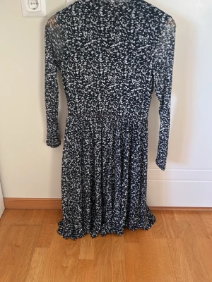 Svart blommig långärmad klänning - Mysig svart klänning med vitt blommönster och lång ärm. Klänningen har en rund halsringning och ett lätt utställt kjolparti som ger en fin siluett. Perfekt för dig som gillar en enkel men ändå söt stil.