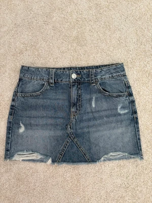 Blå jeanskjol med fickor på baksidan  - Trendig blå jeanskjol i kort modell med slitna detaljer och rå kant nertill. Kjolen har klassiska fickor fram och bak samt knapp och dragkedja framtill. Den är från Gina tricot barn avdelning. Är i mycket bra skick och endast använd ca 2-3 gånger. 