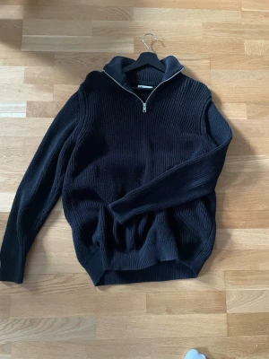 Svart stickad tröja med halv dragkedja - Svart stickad tröja med hög krage och halv dragkedja framtill. Tröjan har ribbstickade muddar vid ärmslut och nederkant, samt en avslappnad passform. Perfekt för lager på lager under kyliga dagar.