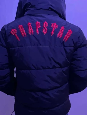 Svart pufferjacka från Trapstar - Säljer en svart pufferjacka från Trapstar med stor röd logotyp broderad på ryggen och en rund patch på bröstet. Jackan har huva, dragkedja framtill och breda kanaler för extra värme. Perfekt för dig som vill ha streetwear-vibe och hålla dig varm.