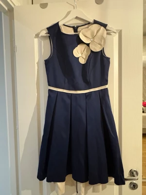 Marinblå kortklänning med blommor - Snygg marinblå klänning från Karen Millen med krämvita detaljer och stora dekorativa blommor vid axeln. Klänningen är ärmlös, har rund hals och ett krämvitt band i midjan. Materialet är glansigt och känns lyxigt, perfekt för dig som vill sticka ut.