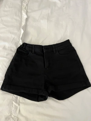 Svarta high-waist shorts i denim - Svarta shorts i denim med hög midja in och uppvikta benslut. Klassisk femficksmodell med bälteshällor och dragkedja framtill. Perfekta för sommaren och enkla att matcha.