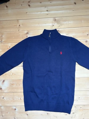 Marinblå stickad tröja Polo Ralph Lauren - Snygg marinblå stickad tröja från Polo Ralph Lauren med halv dragkedja och ribbad krage. Tröjan har det klassiska röda Polo-logot broderat på bröstet och ribbade muddar vid ärmslut och nederkant. Perfekt för lager på lager och stilren look.